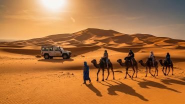 Du khách trên đoàn lạc đà băng qua đồi cát Erg Chebbi gần Merzouga, xe 4x4 đỗ gần đồi cát