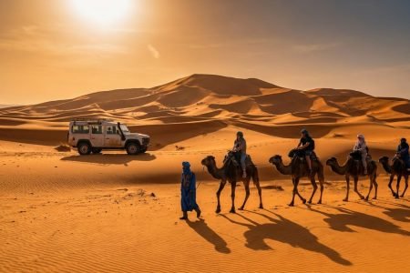 Du khách trên đoàn lạc đà băng qua đồi cát Erg Chebbi gần Merzouga, xe 4x4 đỗ gần đồi cát