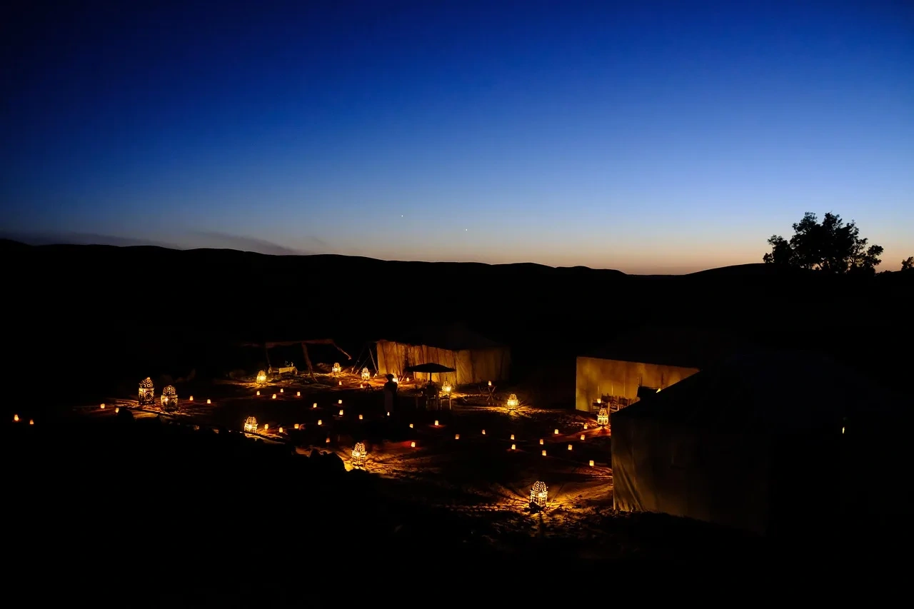 A cluster of tents at a desert camp lit up at dusk in the Sahara.