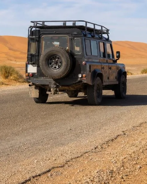 4x4 vehicle with roof rack and spare wheel driving a desert track toward the golden Sahara dunes near Merzouga